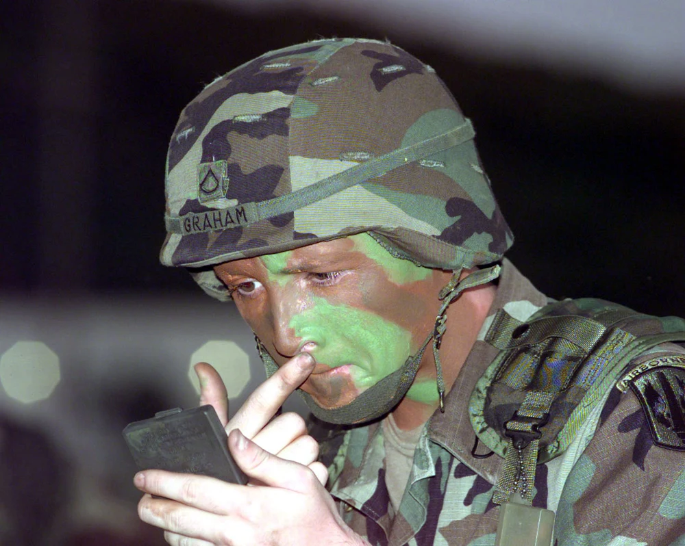 A soldier wearing a patterned helmet applies green camouflage paint to his face.