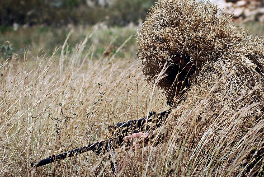 A person in a grass-coloured ghillie suit crouches among tall dry grass, with the head and shoulders covered in loose fibres.