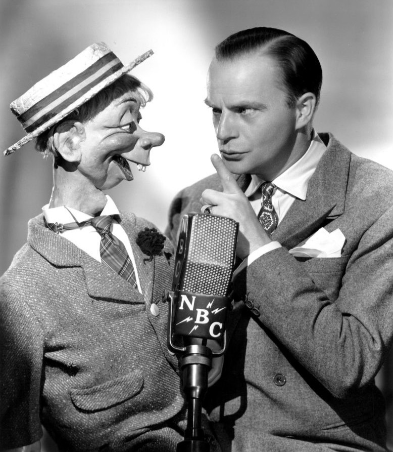 A black-and-white photograph of a ventriloquist speaking beside his puppet.