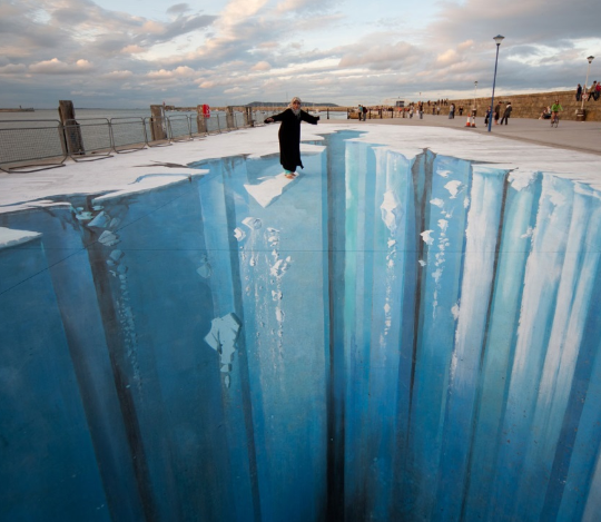 A person stands beside a pavement painting that appears to open into a vast blue crevasse.
